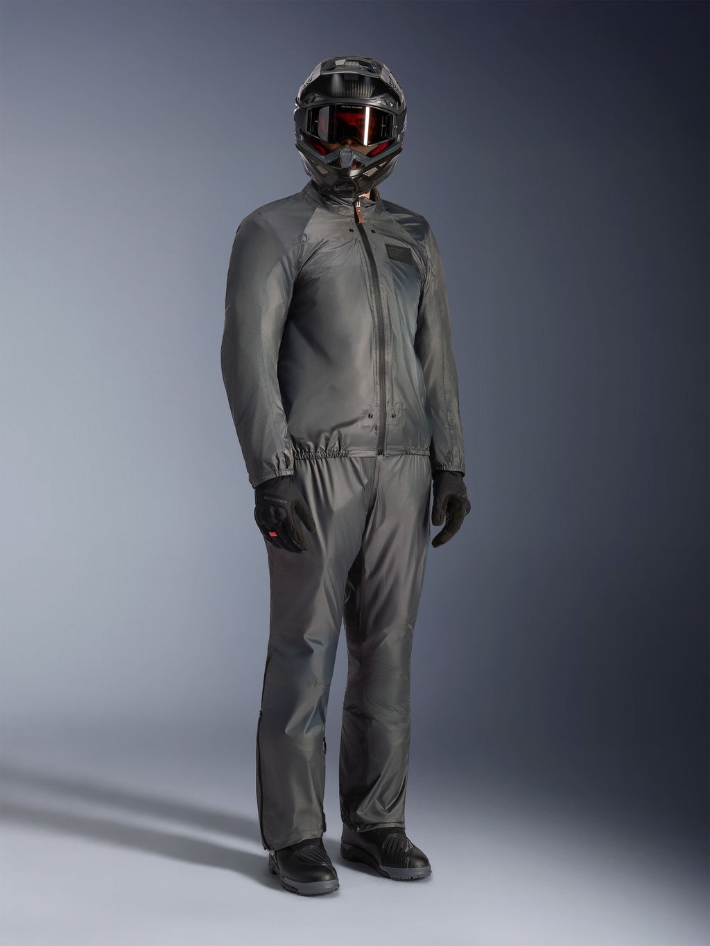 Piloto con la chaqueta Alpinestars Borrego Drystar® en gris cálido, equipamiento de aventura completo con pantalones grises a juego, casco integral negro con gafas y guantes negros, con un elegante diseño textil impermeable.