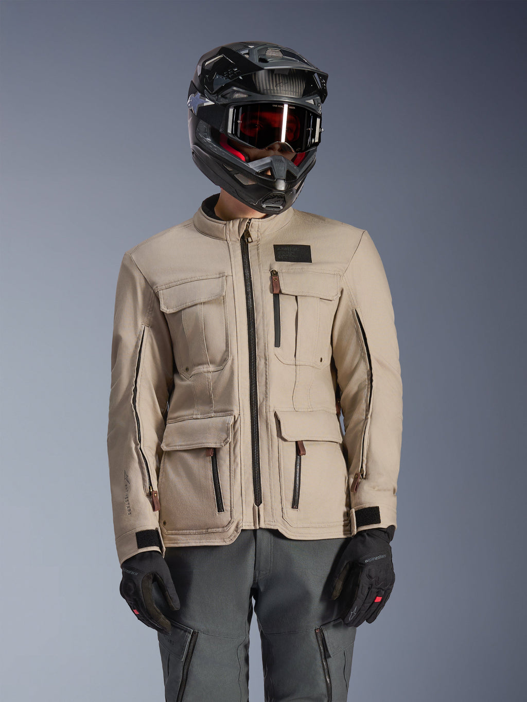 Chaqueta Alpinestars Borrego Drystar®, chaqueta de aventura para moto, color Warm Gray, con múltiples bolsillos cargo frontales y cremalleras negras, combinada con casco negro, gafas y pantalones de moto gris oscuro, diseño textil robusto para touring