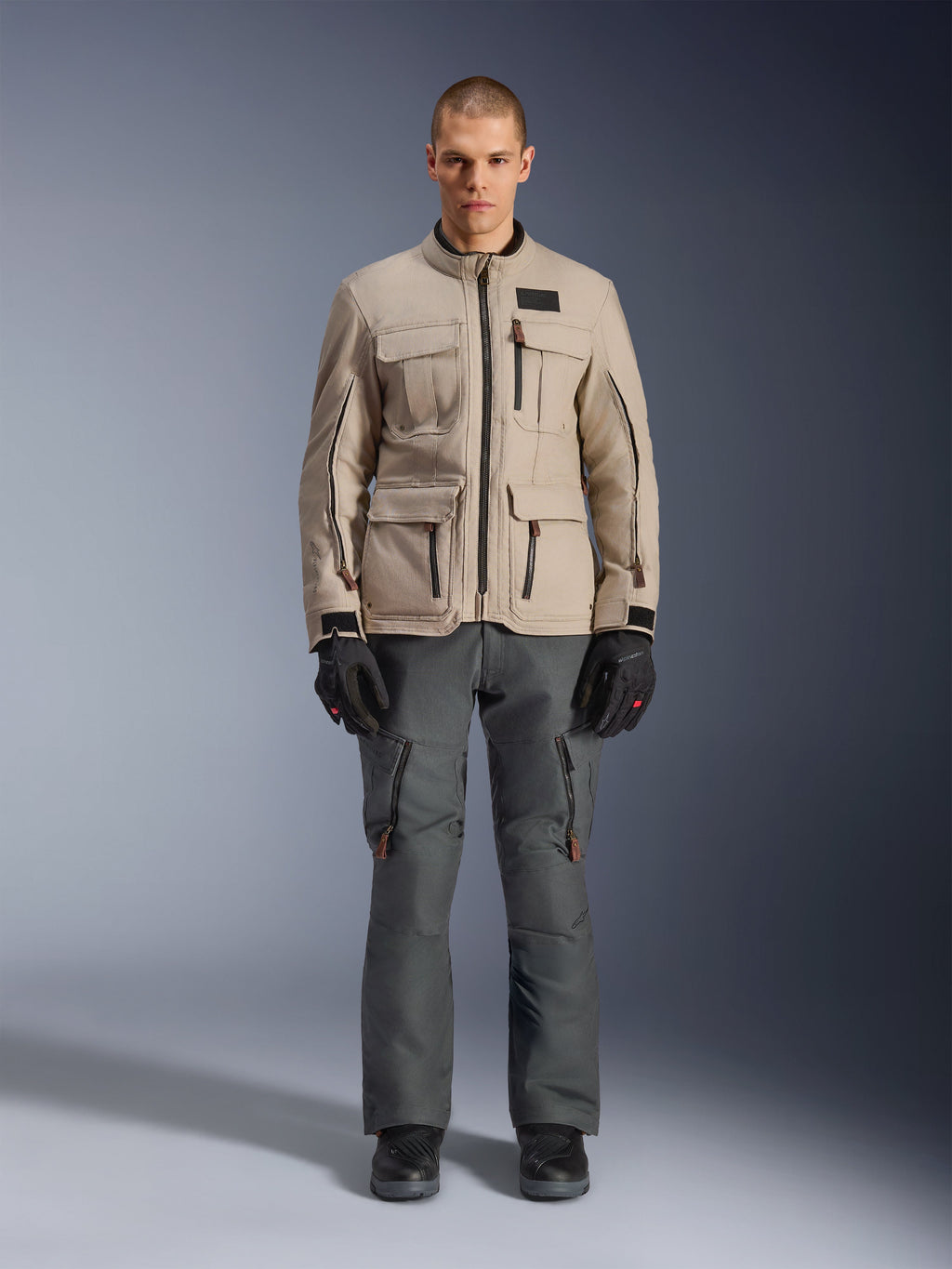 Chaqueta Alpinestars Borrego Drystar®, chaqueta de moto de aventura, gris cálido, vestida por un modelo con pantalones de conducción gris oscuro y guantes negros, con múltiples bolsillos tipo cargo y una construcción textil duradera con un aspecto clásico de lona encerada.