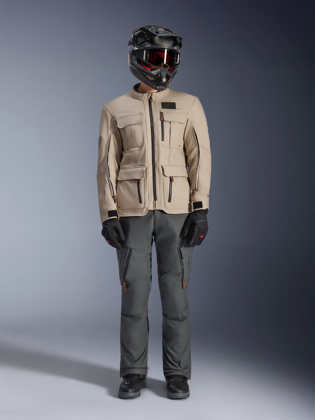 Chaqueta Alpinestars Borrego Drystar® en color Warm Gray, chaqueta de moto de aventura con cuatro bolsillos de carga frontales, combinada con pantalones de moto gris oscuro, casco off-road negro y guantes negros, con un robusto aspecto de lona encerada
