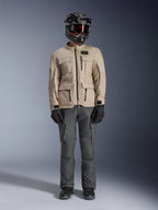 Chaqueta Alpinestars Borrego Drystar® en color Warm Gray, chaqueta de moto de aventura con cuatro bolsillos de carga frontales, combinada con pantalones de moto gris oscuro, casco off-road negro y guantes negros, con un robusto aspecto de lona encerada