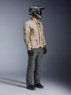 Motociclista con chaqueta Alpinestars Borrego Drystar® en color gris cálido (Warm Gray), chaqueta de moto de aventura con textura de lona encerada y bolsillos utilitarios, combinada con pantalones de conducción gris oscuro, casco de fibra de carbono, guantes y botas negras, vista de estudio de cuerpo completo de un conjunto de touring de aventura