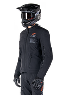 Chaqueta Alpinestars AMT-8 Stretch Drystar® XF, negra, usada por un piloto con pantalones de aventura y guantes a juego, con un casco de motocross negro y gafas, puertos de ventilación con cremallera en el pecho y los brazos, marca Alpinestars Motion Technology con un logotipo naranja, equipamiento premium de turismo de aventura