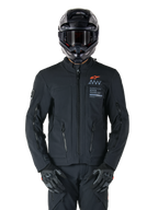 Chaqueta Alpinestars AMT-8 Stretch Drystar® XF, chaqueta de moto negra con logotipo naranja de Alpinestars y marca AMT en gris, cuello alto con cremallera frontal y ventilaciones, combinada con casco y guantes negros, equipamiento de protección para adventure touring