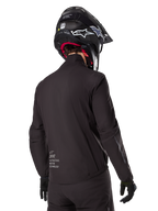 Alpinestars AMT-8 Stretch Drystar® XF Liner, forro de chaqueta impermeable negro, vista trasera de un piloto con casco y guantes negros, con el logotipo Alpinestars Motion Technology en la parte inferior de la espalda, diseño minimalista y funcional para conducción de aventura.