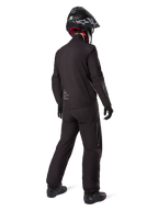 Vista trasera de un motorista que lleva el forro de la chaqueta Alpinestars AMT-8 Stretch Drystar® XF, equipamiento técnico de moto en color negro, combinado con pantalones a juego y casco negro, con el logotipo "AMT Alpinestars Motion Technology" en la espalda, diseño minimalista y elegante para touring de aventura