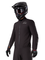 Piloto vistiendo el forro Alpinestars AMT-8 Stretch Drystar® XF, chaqueta elástica negra cortavientos e impermeable con logotipo AMT en el pecho y marca Drystar XF en la manga, combinada con casco de aventura negro y guantes protectores.
