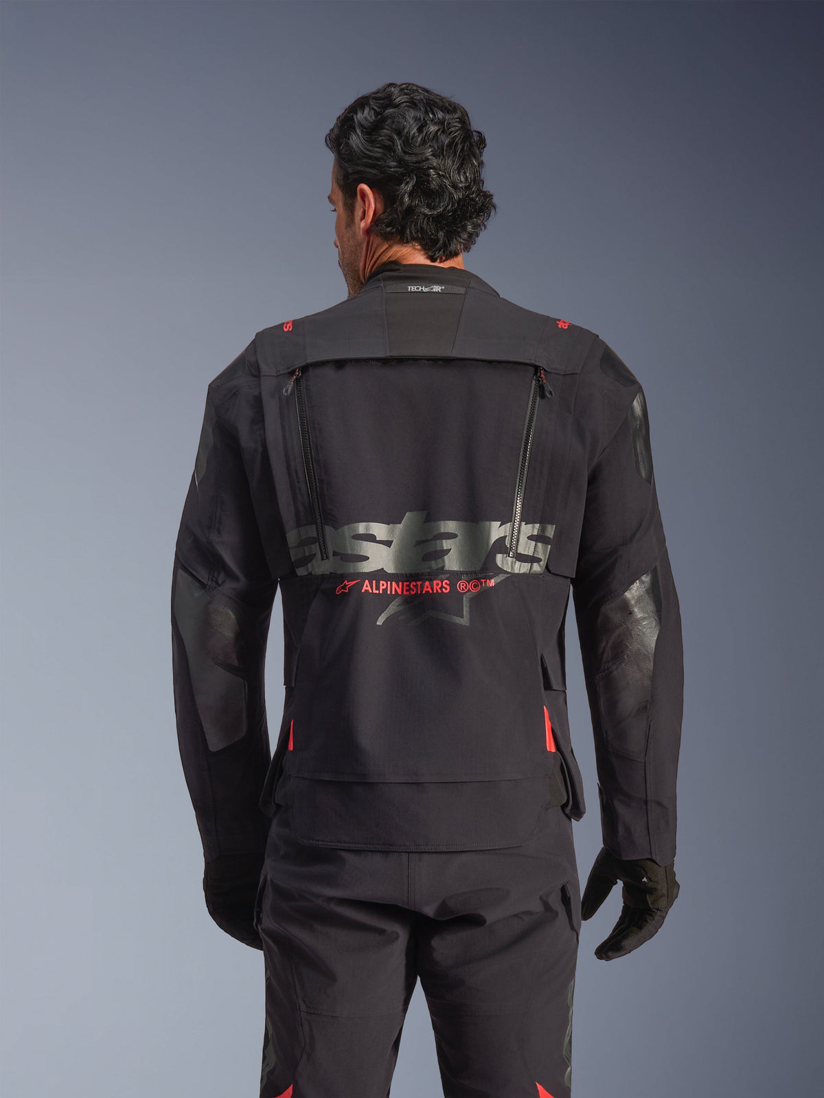 Chaqueta laminada Alpinestars Halo Pro Drystar® XF en Black Dark Shadow, vista trasera en modelo, que muestra el panel técnico de ventilación posterior con cremalleras, detalles del logo de Alpinestars en rojo y marca Tech-Air® en el cuello, equipamiento modular para conducción de aventura.