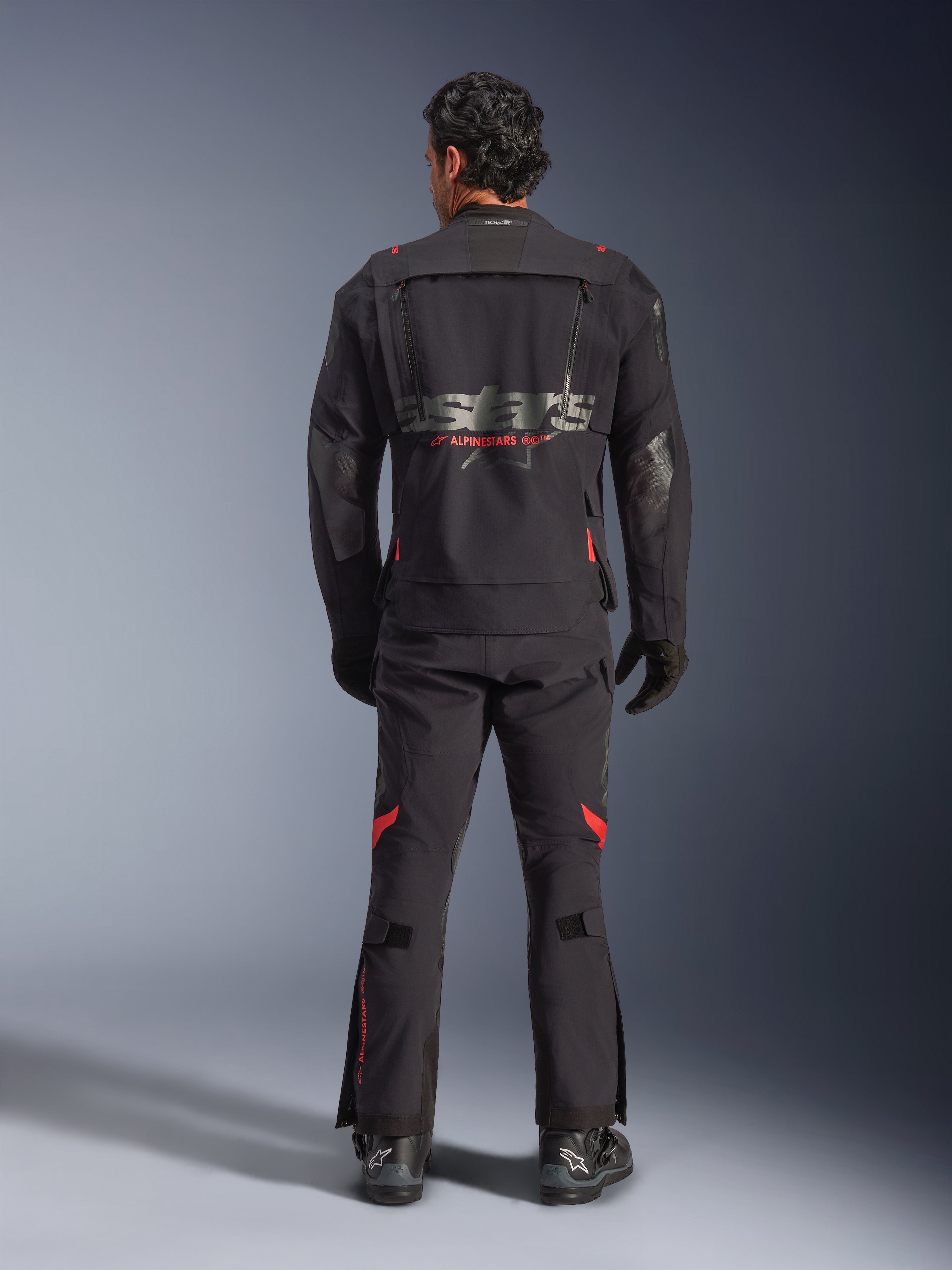 Un piloto visto desde atrás vistiendo la chaqueta laminada Alpinestars Halo Pro Drystar® XF en Black Dark Shadow, con un gran logotipo astars gris y marca Alpinestars en rojo, paneles de ventilación modulares en la espalda, combinada con pantalones de aventura a juego y botas negras.