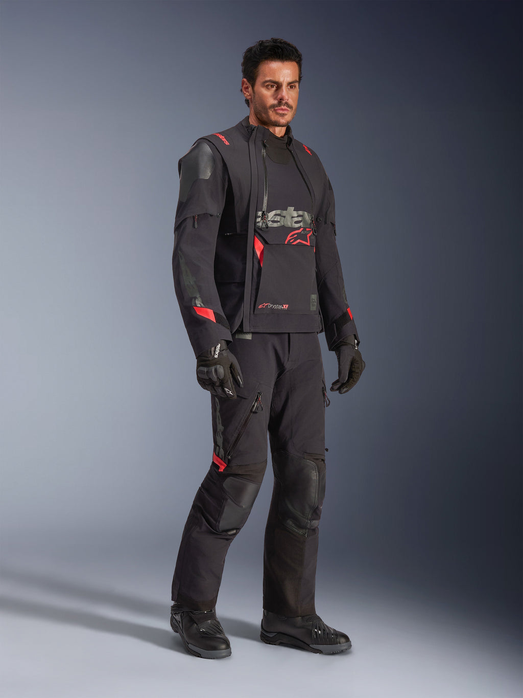 Piloto con la chaqueta laminada Alpinestars Halo Pro Drystar® XF en Black Dark Shadow, equipamiento de aventura completo con pantalones, guantes y botas a juego, paneles frontales asimétricos modulares y detalles rojos de la marca.