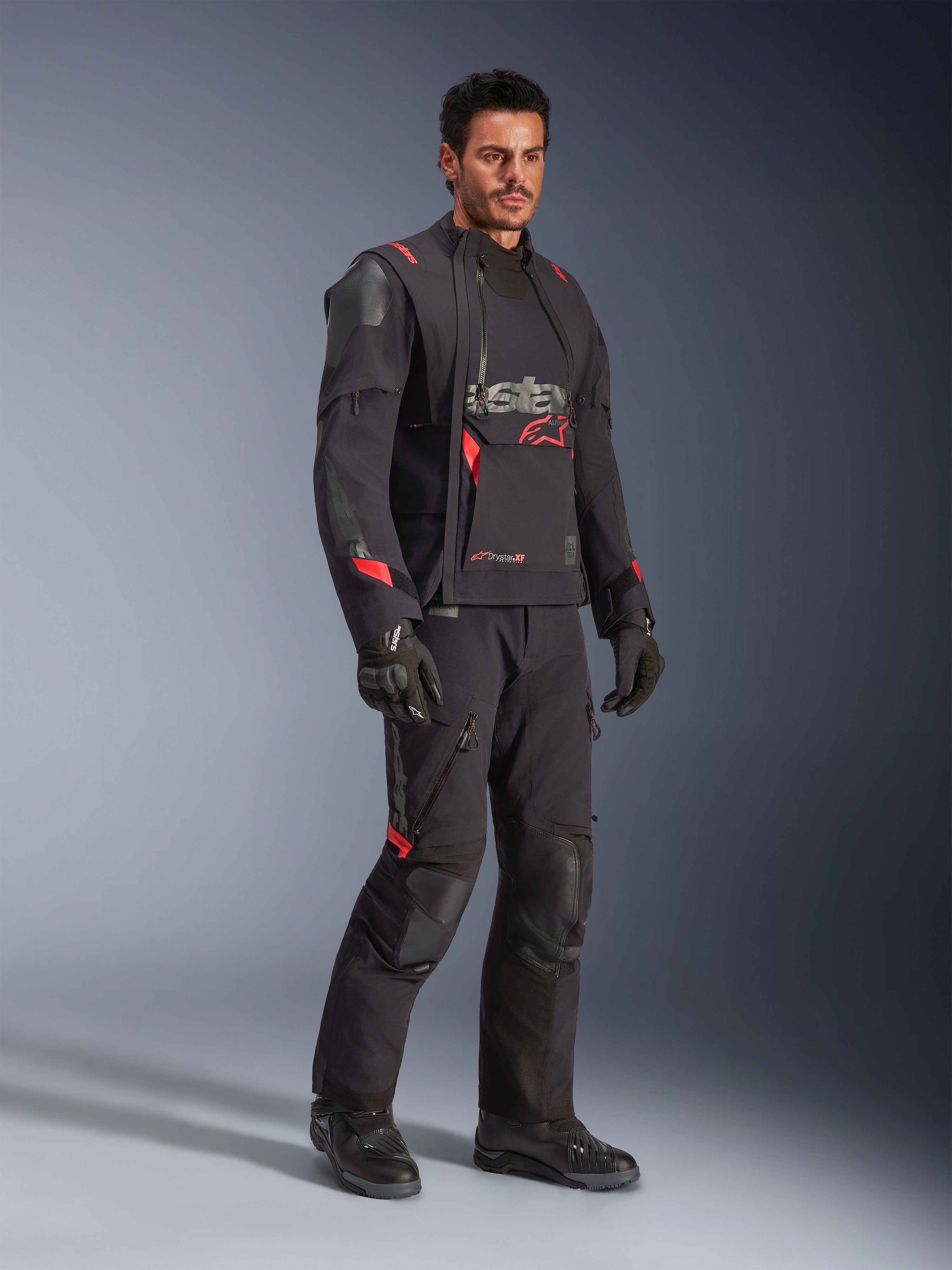 Piloto con la chaqueta laminada Alpinestars Halo Pro Drystar® XF en Black Dark Shadow, equipamiento de aventura completo con pantalones, guantes y botas a juego, paneles frontales asimétricos modulares y detalles rojos de la marca.
