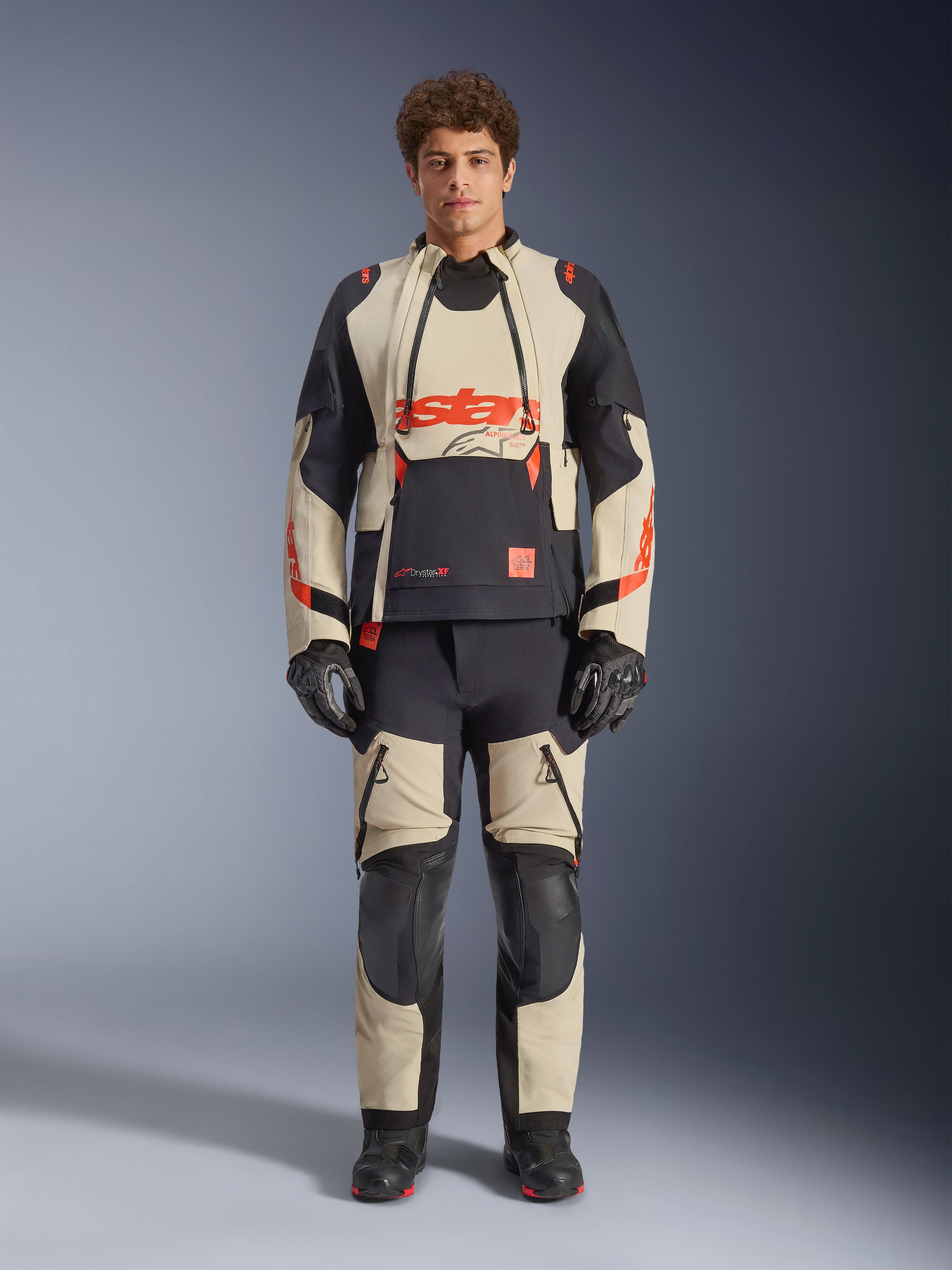 Chaqueta laminada Alpinestars Halo Pro Drystar® XF, Mastic Black, chaqueta de moto de aventura usada por un modelo con pantalones y guantes a juego, paneles textiles en color arena y negro con logotipos rojos llamativos, diseño modular para conducción todoterreno