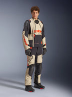 Modelo masculino vistiendo la chaqueta laminada Alpinestars Halo Pro Drystar® XF en color Mastic Black, con paneles beige y negros y detalles en rojo, combinada con pantalones de aventura a juego y guantes negros, diseñada para el motociclismo de aventura todoterreno.