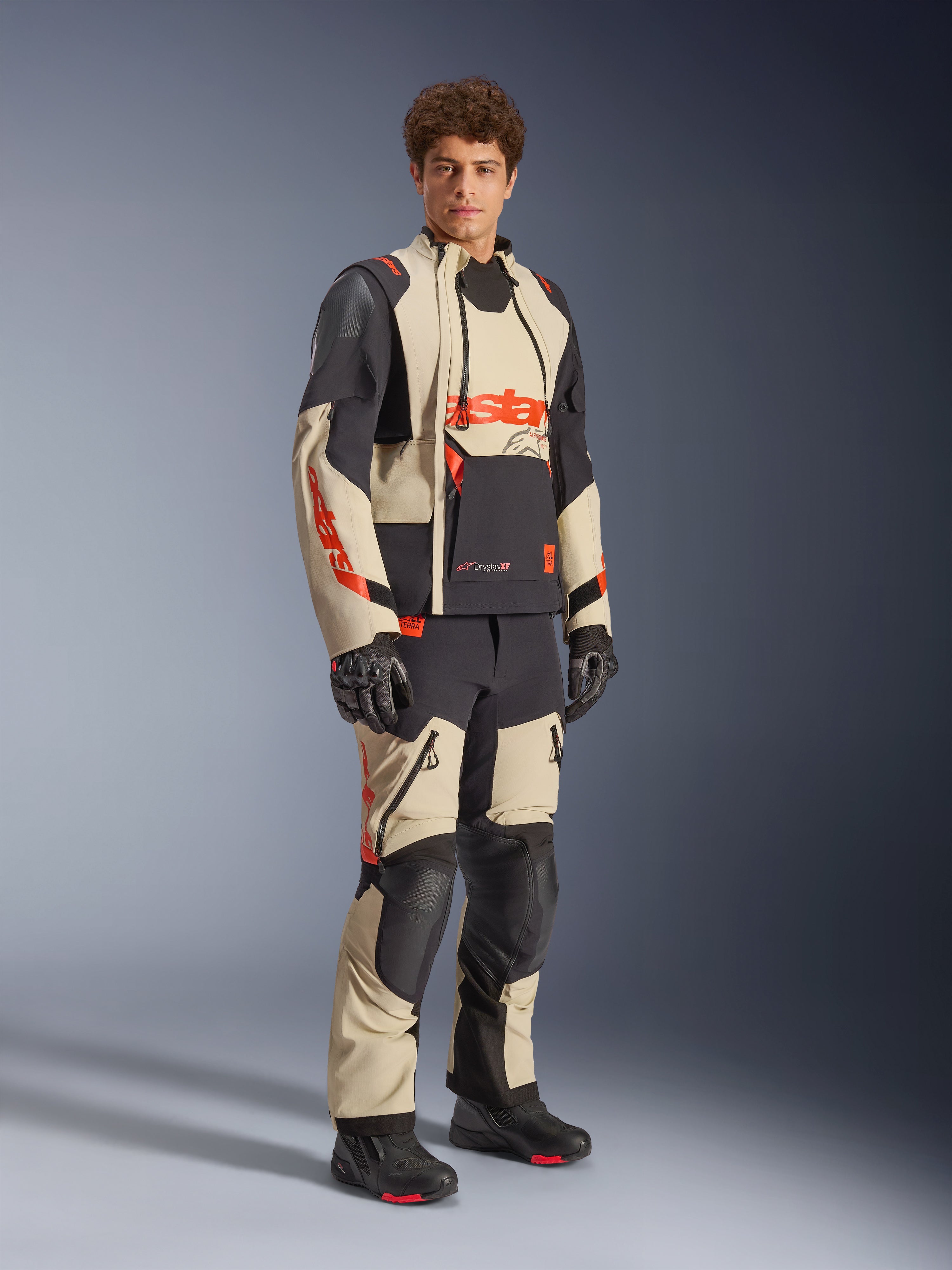 Modelo masculino vistiendo la chaqueta laminada Alpinestars Halo Pro Drystar® XF en color Mastic Black, con paneles beige y negros y detalles en rojo, combinada con pantalones de aventura a juego y guantes negros, diseñada para el motociclismo de aventura todoterreno.