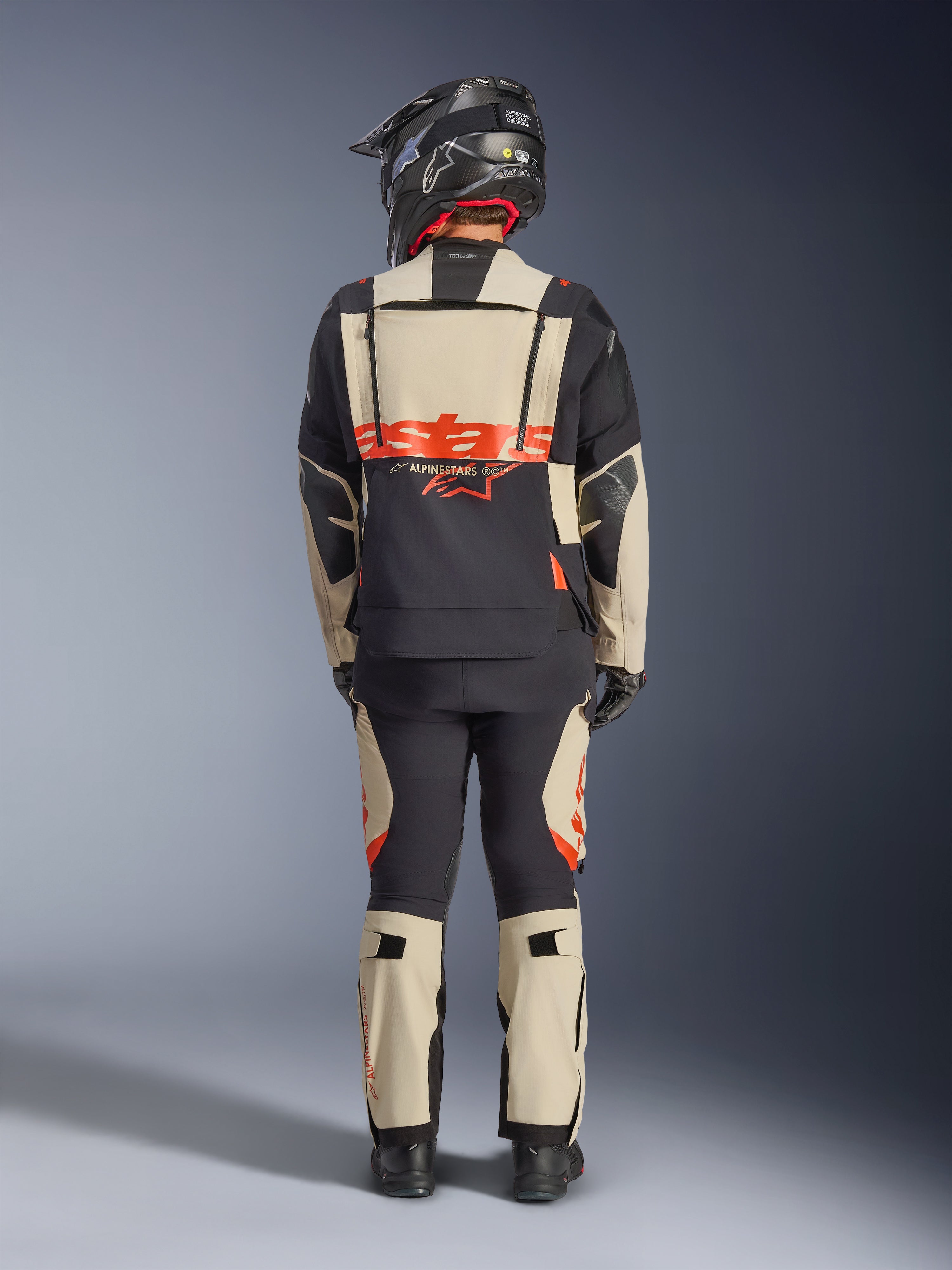 Chaqueta laminada Alpinestars Halo Pro Drystar® XF, chaqueta de aventura para moto, Mastic Black, vista trasera de un piloto con pantalones a juego y un casco negro, con llamativa marca Alpinestars en naranja y paneles de ventilación modulares para una conducción de aventura versátil.