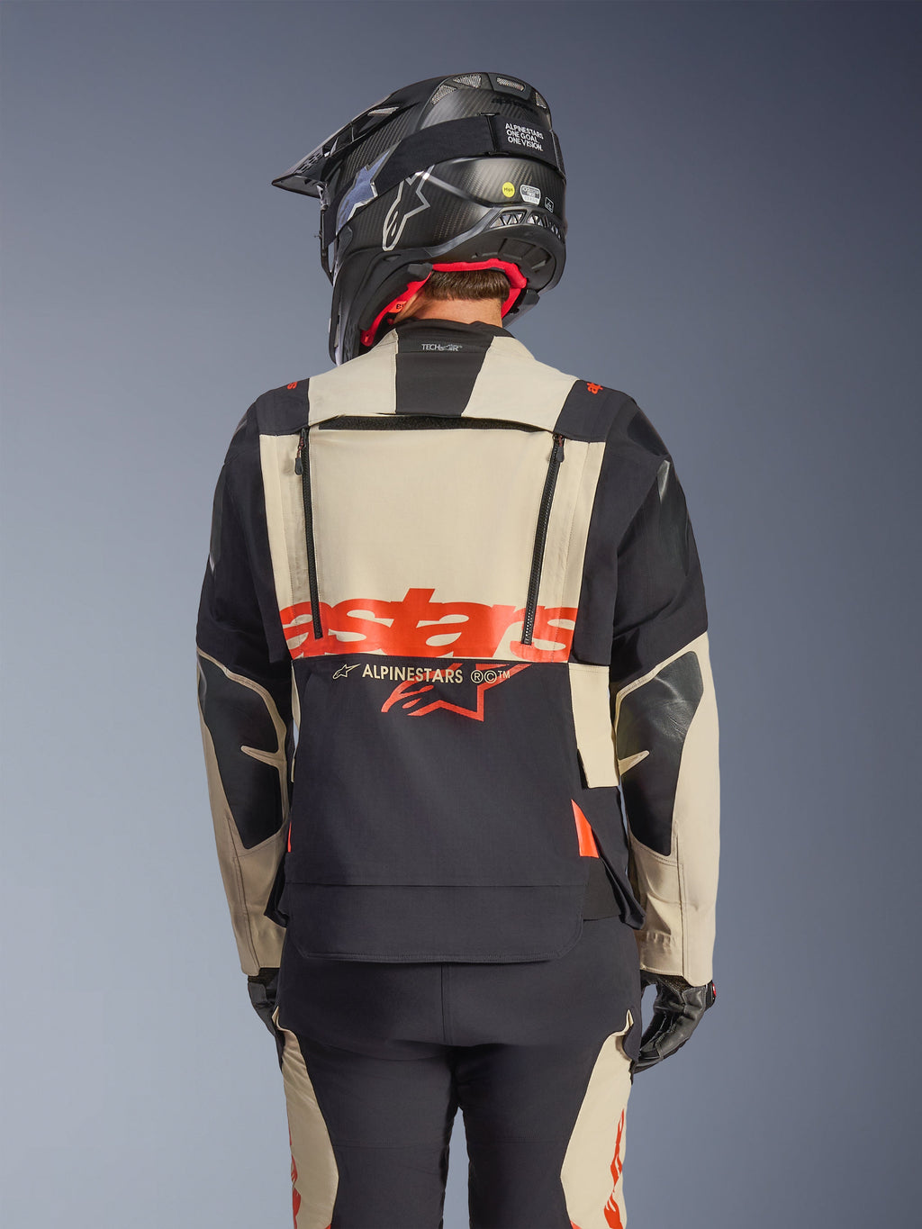 Chaqueta laminada Alpinestars Halo Pro Drystar® XF en Mastic Black, vista trasera que muestra el panel posterior modular con logotipo rojo y cremalleras de ventilación, usada por un piloto con pantalones de aventura a juego y un casco negro