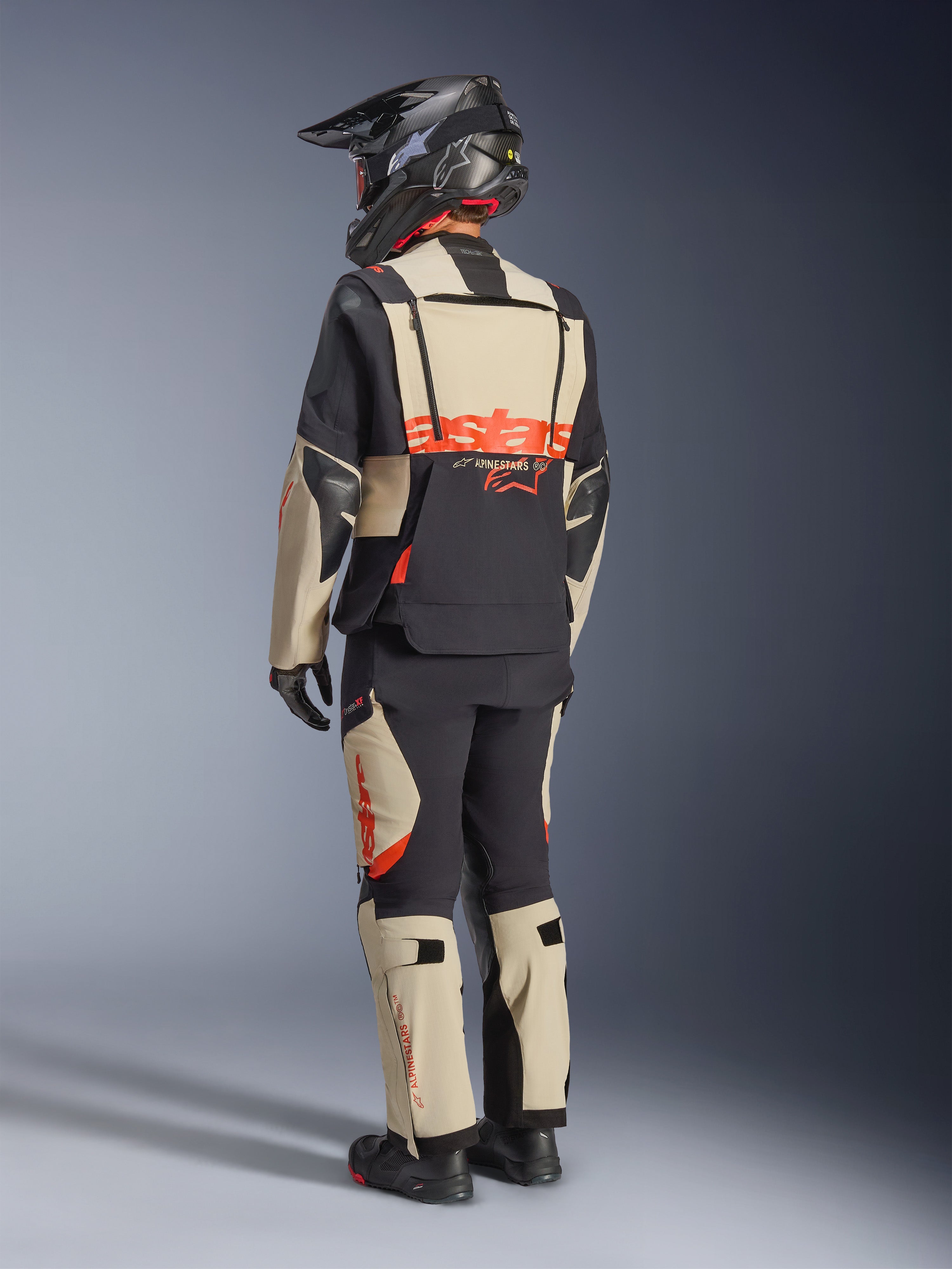 Chaqueta laminada Alpinestars Halo Pro Drystar® XF, Mastic Black, vista trasera de un piloto con el traje de aventura completo, pantalones a juego, casco de aventura negro y botas, con un llamativo logotipo rojo de Alpinestars en la espalda.