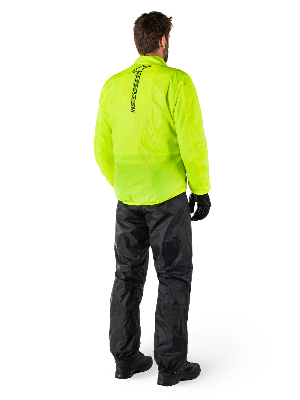 Persona desde atrás con la chaqueta Alpinestars Hurricane Rain V2 en amarillo flúor y el pantalón Hurricane Rain V2 en negro, logotipo vertical en la espalda de la chaqueta, equipo de lluvia impermeable para motocicleta con guantes y botas negros