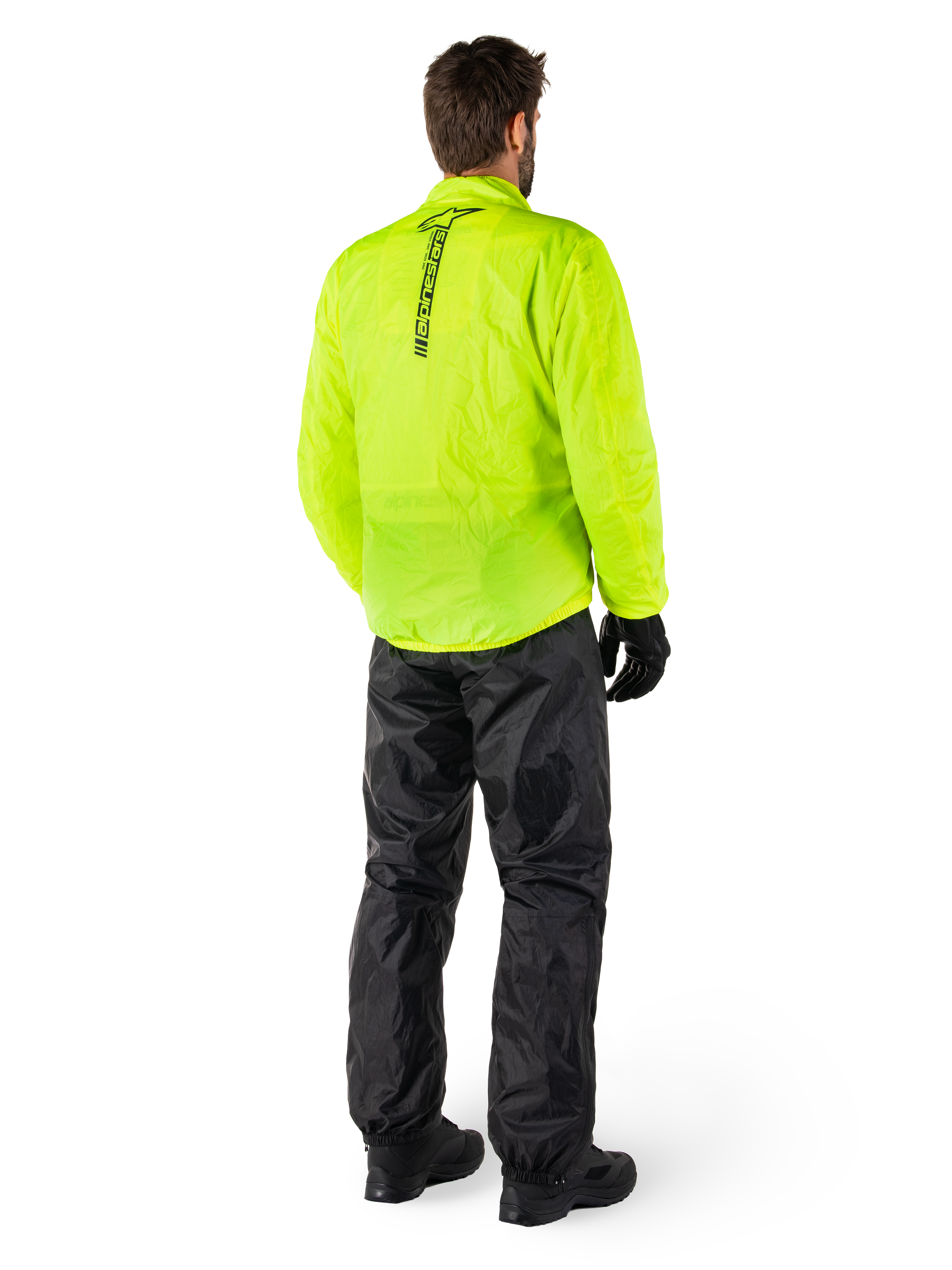 Persona desde atrás con la chaqueta Alpinestars Hurricane Rain V2 en amarillo flúor y el pantalón Hurricane Rain V2 en negro, logotipo vertical en la espalda de la chaqueta, equipo de lluvia impermeable para motocicleta con guantes y botas negros