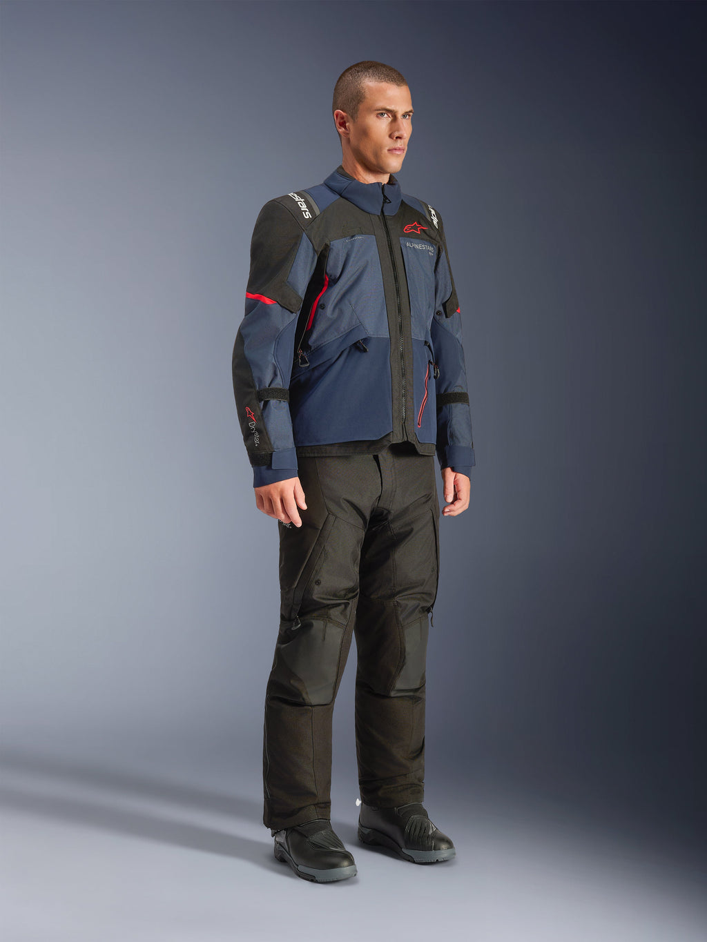 Modelo masculino luciendo la chaqueta Alpinestars Andes V4 Drystar® en Night Navy y Mood Indigo con paneles negros y detalles rojos, combinada con pantalones y botas de moto negros, equipamiento técnico de aventura y touring en un entorno de estudio.