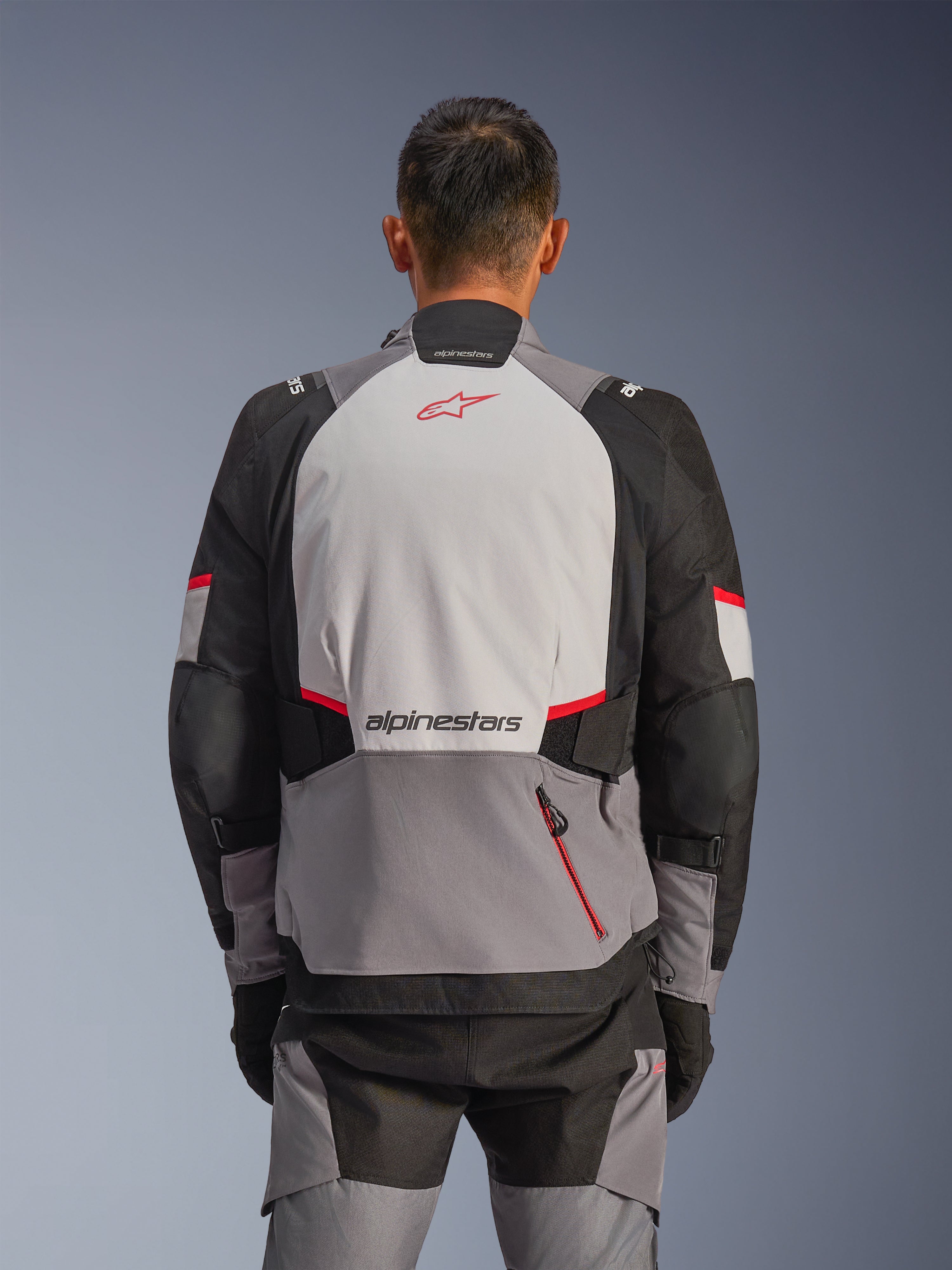 Chaqueta Alpinestars Andes V4 Drystar®, vista posterior, gris oscuro, gris hielo, negro y rojo brillante, con logotipo Astar rojo y marca Alpinestars en la espalda, paneles reforzados y bolsillo con cremallera roja, equipamiento de moto de aventura