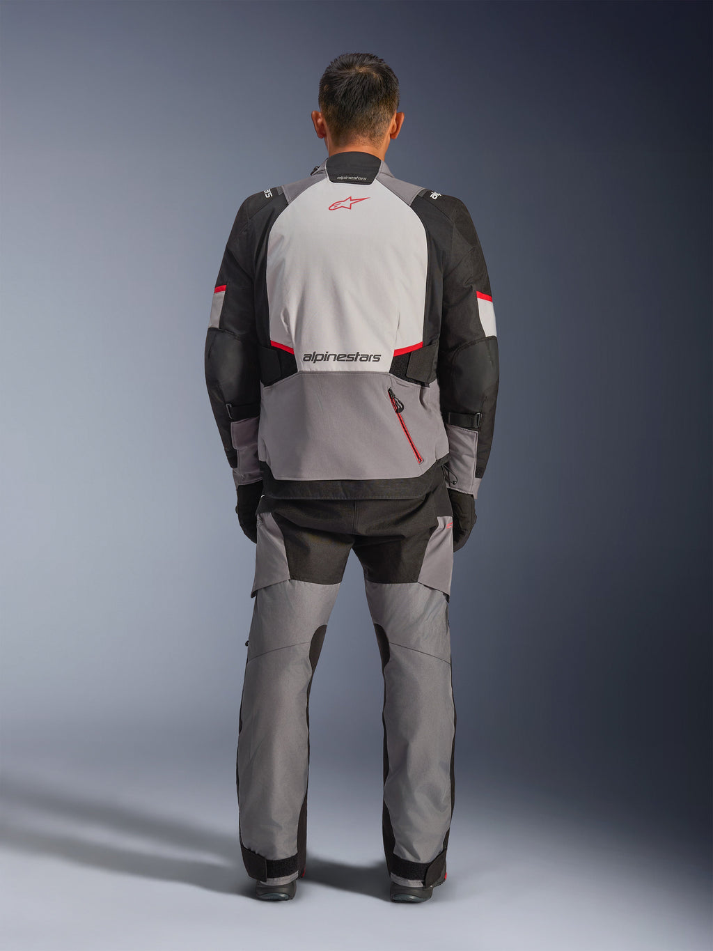 Chaqueta Alpinestars Andes V4 Drystar®, chaqueta de moto de aventura, gris oscuro, gris hielo, negro y rojo brillante, vista trasera de un motorista con pantalones a juego, con un gran panel posterior gris hielo con logotipo rojo, secciones de hombros reforzadas en negro y correas de cintura ajustables para turismo en cualquier clima.