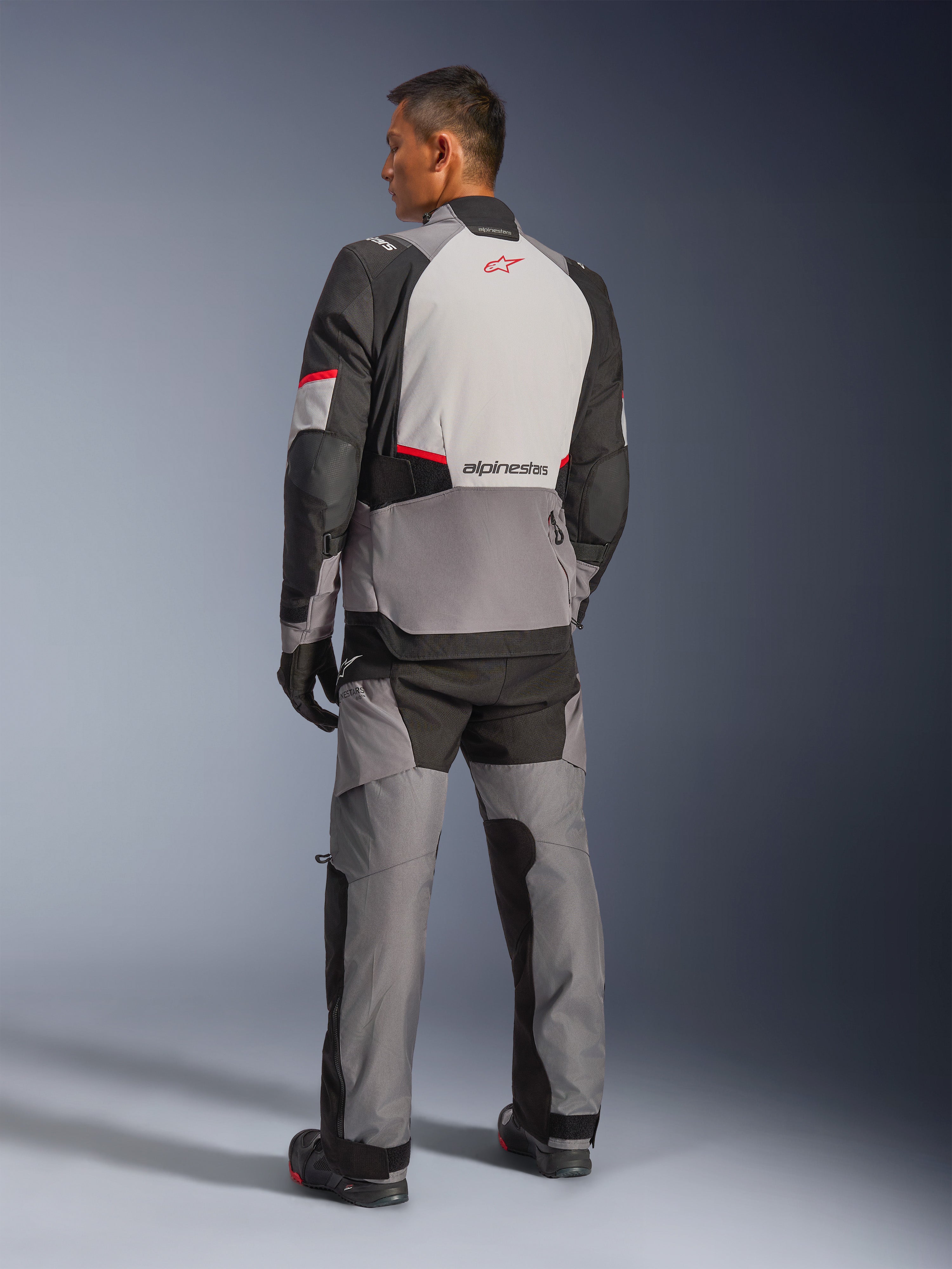 Chaqueta Alpinestars Andes V4 Drystar®, gris oscuro y gris hielo con detalles en negro y rojo brillante, vista trasera con panel gris claro y logo de estrella roja, combinada con pantalones de aventura a juego y guantes negros, equipamiento técnico de motociclismo para todo tipo de clima.