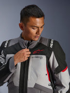 Modelo con la chaqueta Alpinestars Andes V4 Drystar®, gris oscuro y gris hielo con detalles en negro y rojo brillante, ajustando la cremallera frontal, con el logotipo rojo de Astars y la marca Drystar®, estilo adventure touring
