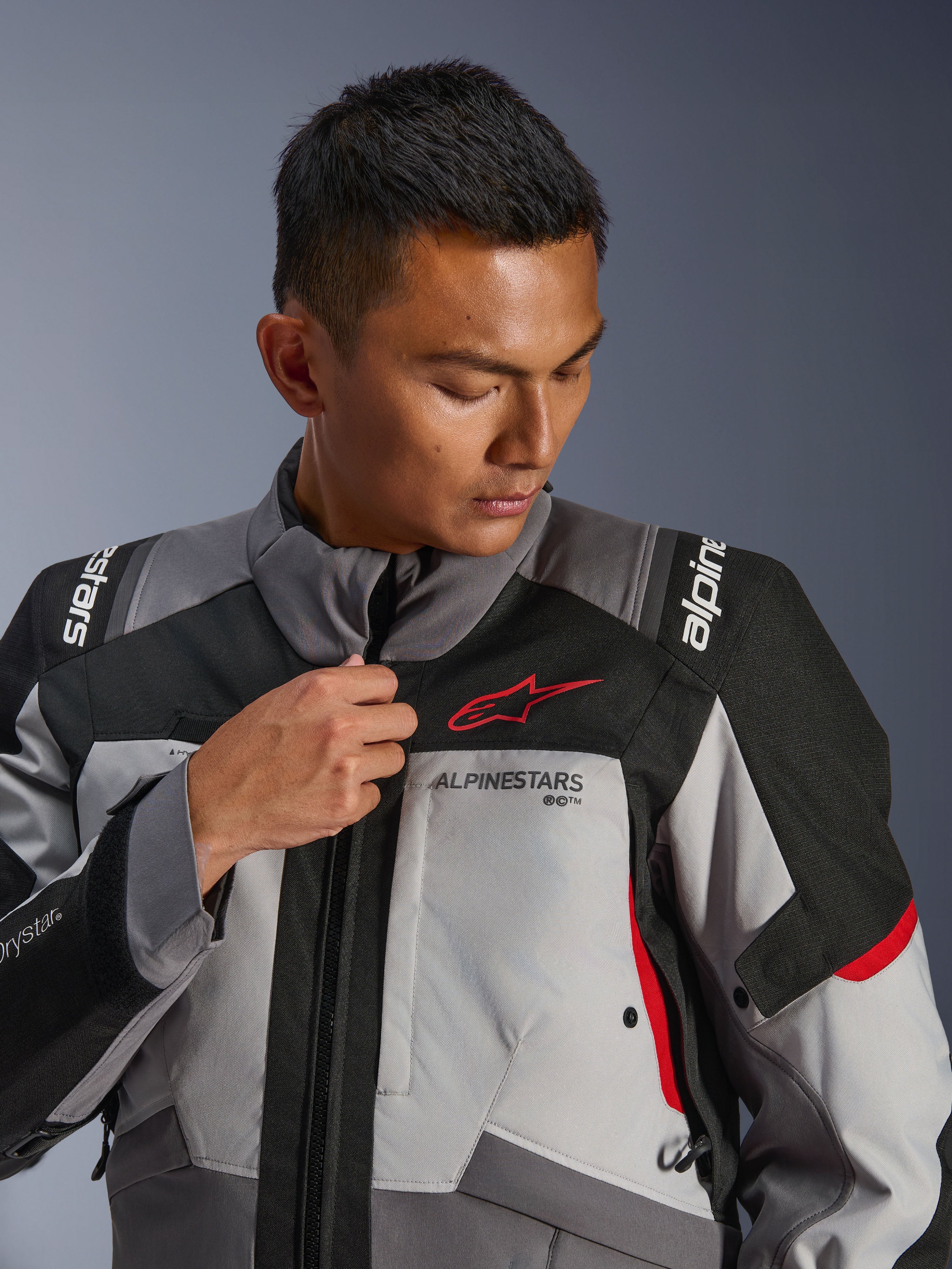 Modelo con la chaqueta Alpinestars Andes V4 Drystar®, gris oscuro y gris hielo con detalles en negro y rojo brillante, ajustando la cremallera frontal, con el logotipo rojo de Astars y la marca Drystar®, estilo adventure touring