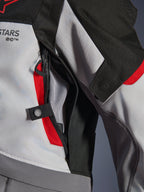 Detalle de primer plano de la chaqueta Alpinestars Andes V4 Drystar® en gris oscuro, gris hielo y rojo brillante, que destaca una abertura de ventilación con cremallera en el pecho con forro de malla y una correa de ajuste con botón a presión.