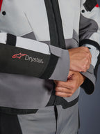 Detalle de primer plano de la manga de la chaqueta Alpinestars Andes V4 Drystar®, textil gris y negro, con el logotipo Drystar en rojo y blanco en el antebrazo, cierre de velcro ajustable en la muñeca, equipamiento impermeable de alto rendimiento para moto