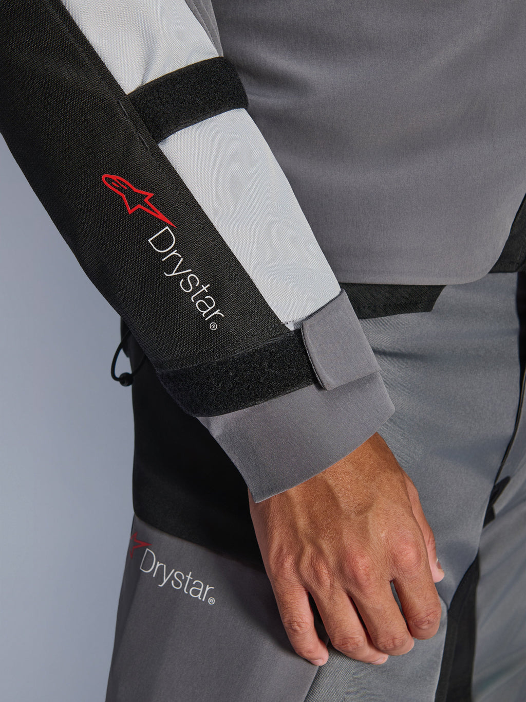 Primer plano de la manga de la chaqueta Alpinestars Andes V4 Drystar®, gris oscuro y gris hielo con detalles en negro, con el logotipo Drystar® y correas ajustables en el antebrazo, parte de un conjunto textil impermeable para moto de aventura