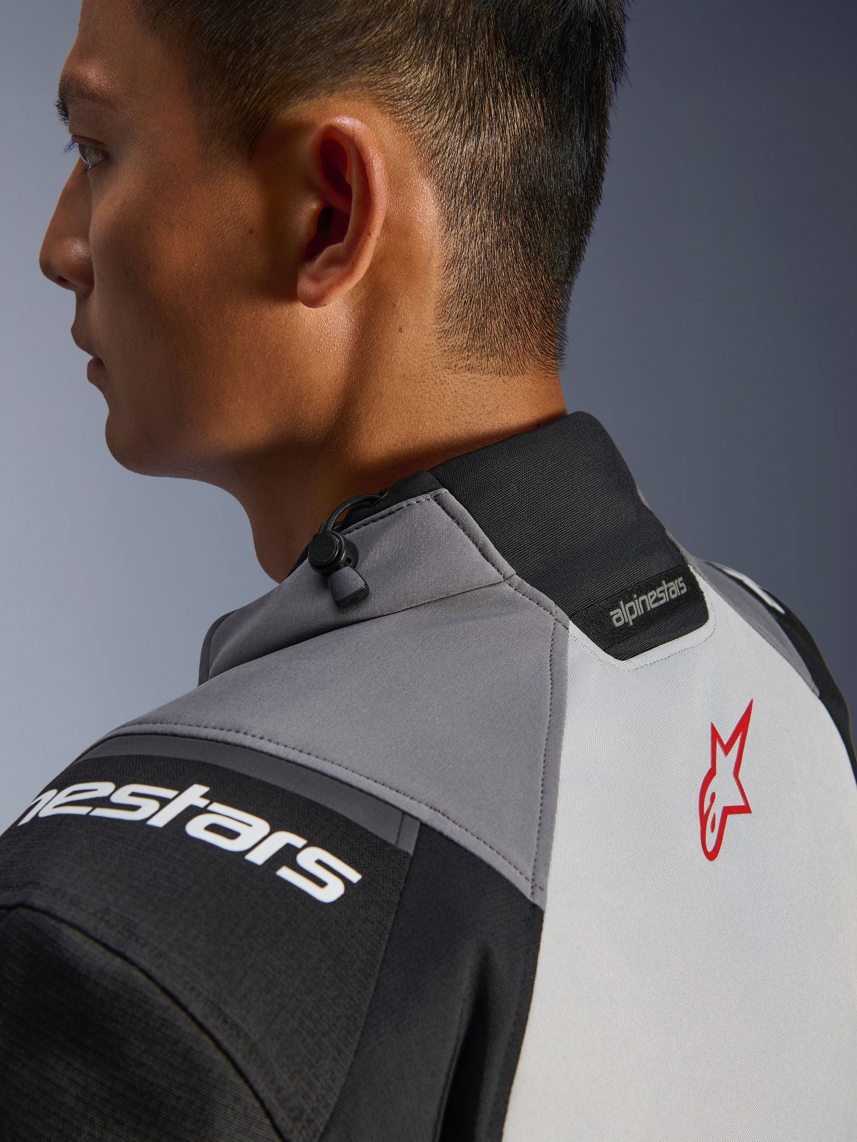 Chaqueta Alpinestars Andes V4 Drystar®, primer plano del cuello y la parte superior de la espalda, gris oscuro y gris hielo con acentos negros, logotipo de estrella roja, con ajustador de cordón en el cuello y marca Alpinestars, construcción textil duradera