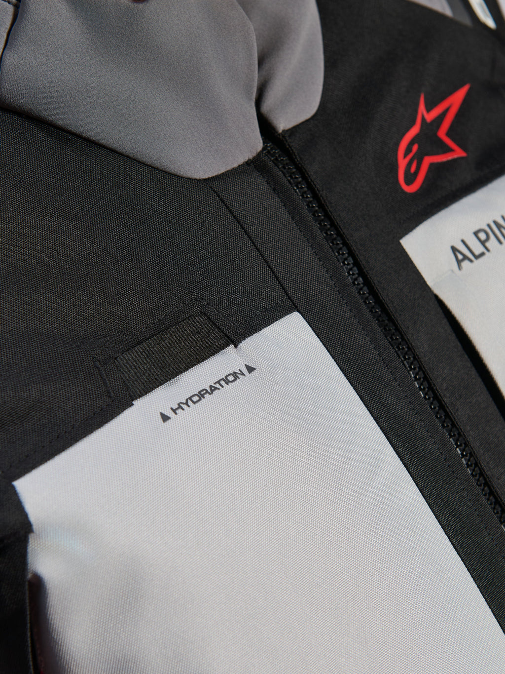 Chaqueta Alpinestars Andes V4 Drystar®, detalle en primer plano del pecho, paneles en gris oscuro y gris hielo con refuerzos textiles negros, con el logotipo de Alpinestars en rojo y un bucle para la manguera de hidratación con la etiqueta 'HYDRATION'