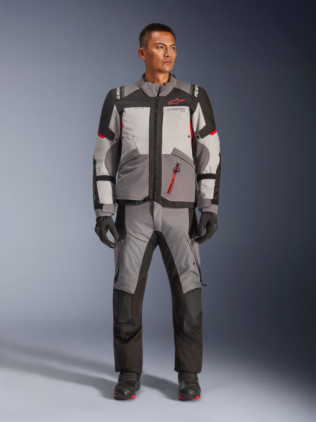 Chaqueta Alpinestars Andes V4 Drystar®, chaqueta de aventura para motocicleta, en gris oscuro, gris hielo, negro y rojo brillante, combinada con pantalones y guantes textiles de conducción a juego, con un diseño técnico de múltiples paneles y el logotipo de Alpinestars en rojo, equipamiento adventure-touring.