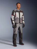 Modelo masculino vistiendo la chaqueta Alpinestars Andes V4 Drystar® y pantalones de aventura a juego, en gris oscuro, gris hielo, negro y rojo brillante, combinados con guantes y botas de moto negros, equipamiento técnico de conducción para todo clima con un moderno diseño multipanel.