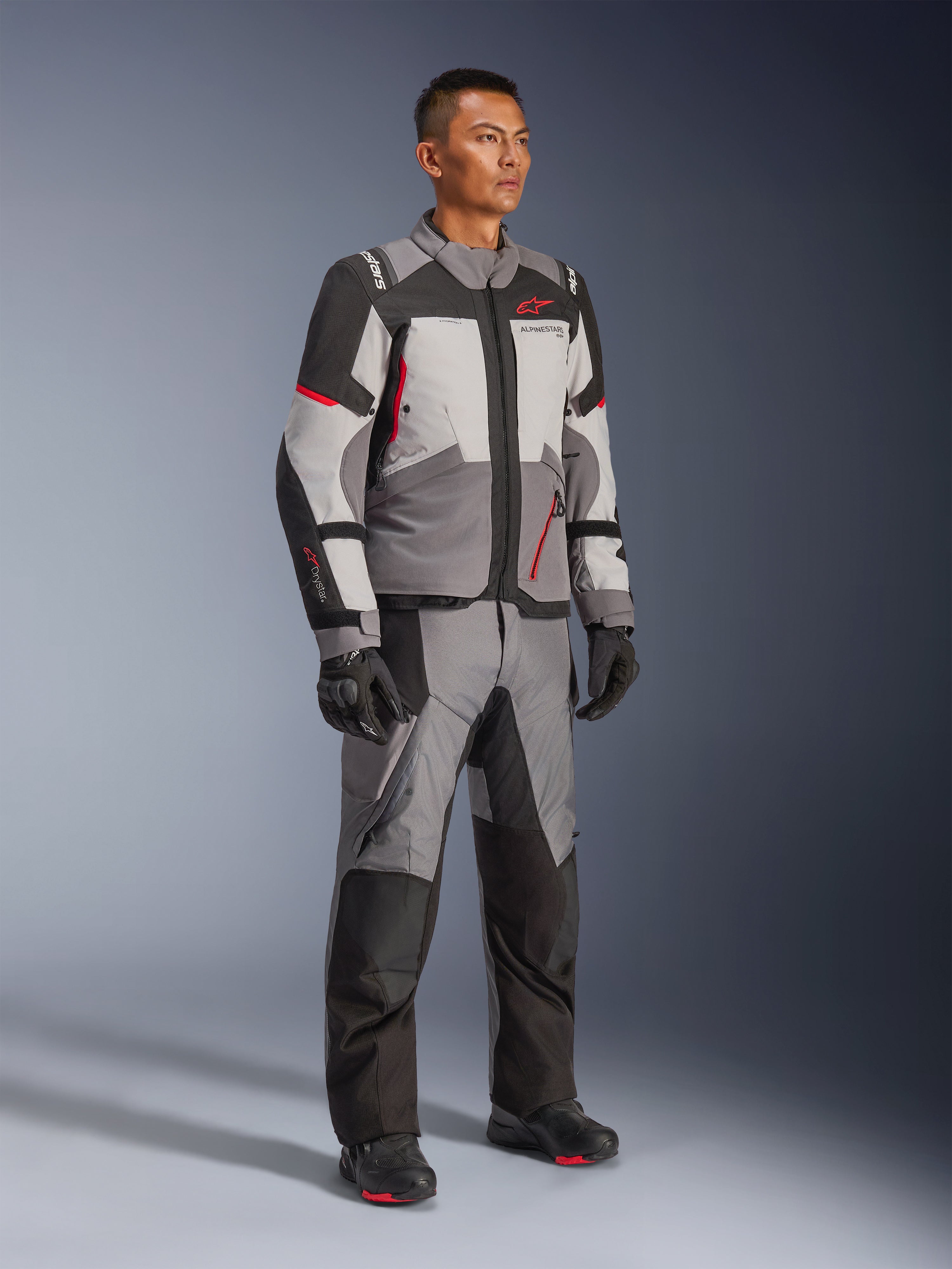 Modelo masculino vistiendo la chaqueta Alpinestars Andes V4 Drystar® y pantalones de aventura a juego, en gris oscuro, gris hielo, negro y rojo brillante, combinados con guantes y botas de moto negros, equipamiento técnico de conducción para todo clima con un moderno diseño multipanel.
