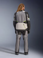 Chaqueta Alpinestars Andes V4 Drystar®, chaqueta de moto de aventura, gris oscuro y gris hielo con detalles en negro y amarillo flúor, vista trasera usada con pantalones a juego y guantes negros, con un gran bolsillo trasero con cremallera amarilla y paneles reforzados en hombros y codos.