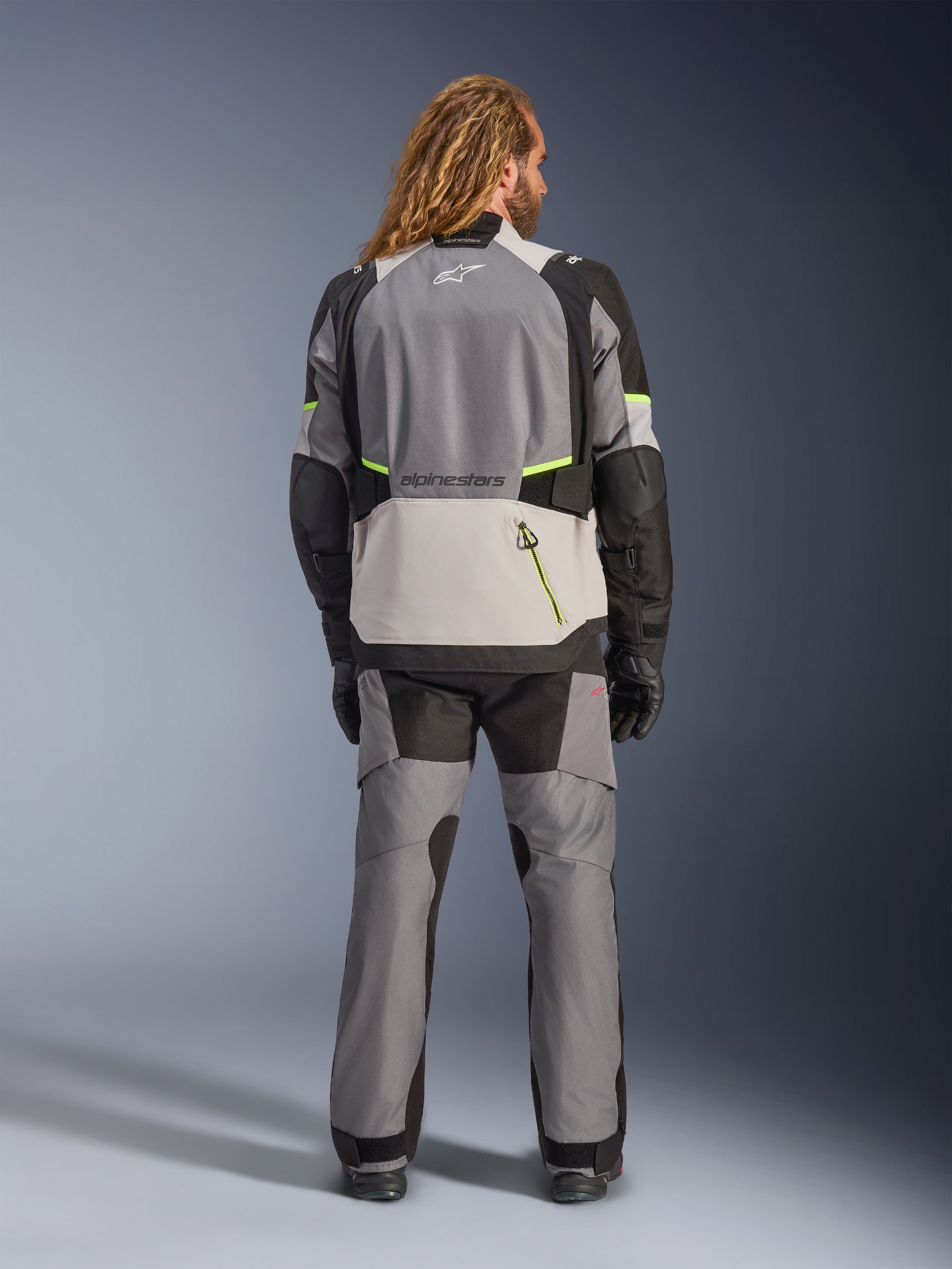 Chaqueta Alpinestars Andes V4 Drystar®, chaqueta de moto de aventura, gris oscuro y gris hielo con detalles en negro y amarillo flúor, vista trasera usada con pantalones a juego y guantes negros, con un gran bolsillo trasero con cremallera amarilla y paneles reforzados en hombros y codos.