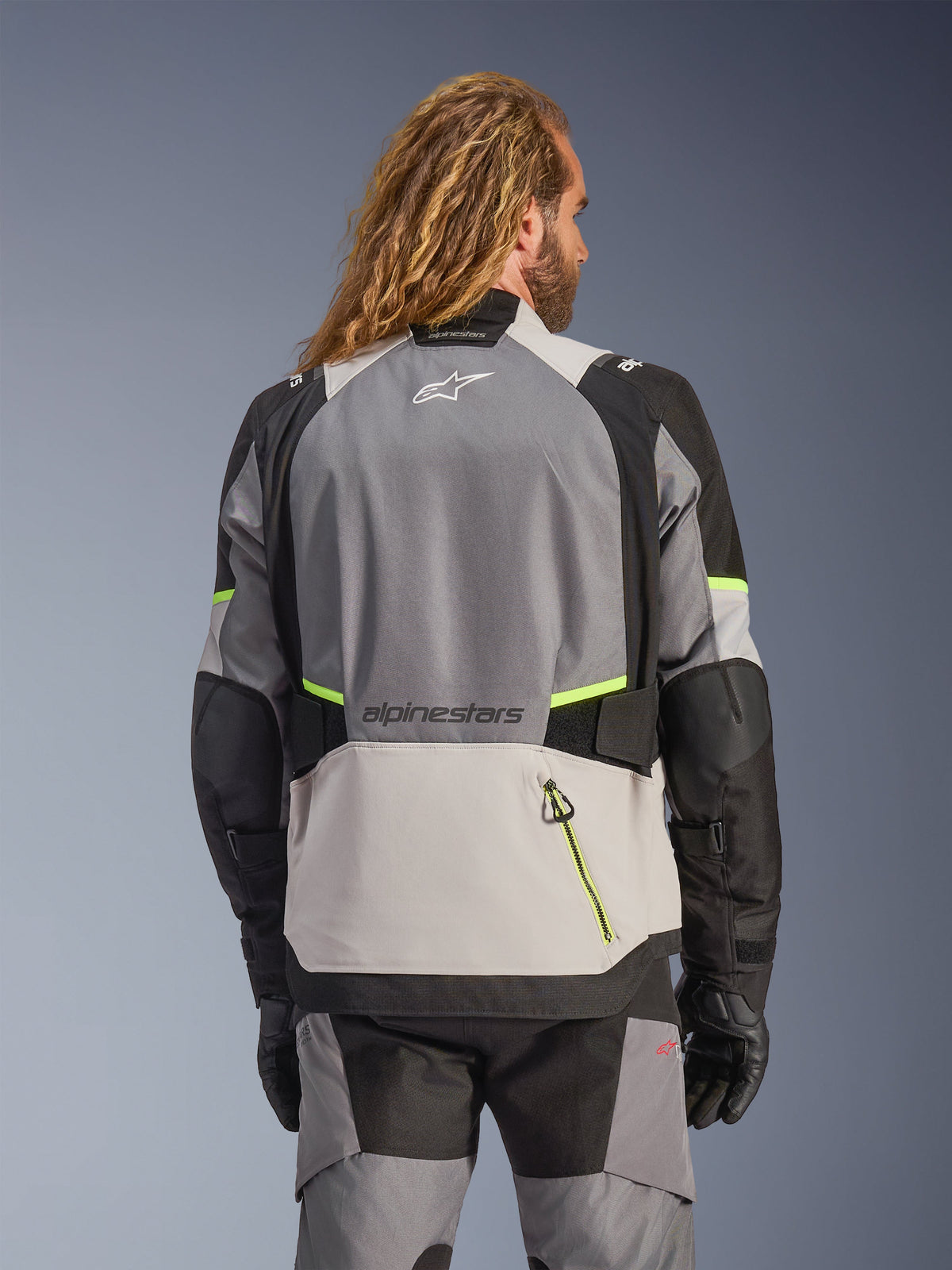 Alpinestars Andes V4 Drystar® Jacket, vista trasera de chaqueta de moto de aventura en gris oscuro, gris hielo y negro con detalles en amarillo flúor, paneles textiles reforzados y logotipos de Alpinestars, usada con pantalones de conducción a juego y guantes negros