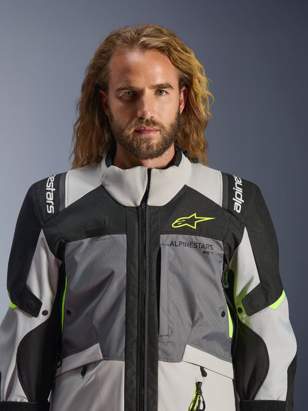 Chaqueta Alpinestars Andes V4 Drystar®, gris oscuro, gris hielo, negro y amarillo flúor, usada por un hombre con cabello largo ondulado, con construcción técnica multitejido, logotipos de Alpinestars en el pecho y los hombros, y diseño de cuello alto para conducción de aventura