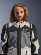 Chaqueta Alpinestars Andes V4 Drystar®, gris oscuro, gris hielo, negro y amarillo flúor, usada por un hombre con cabello largo ondulado, con construcción técnica multitejido, logotipos de Alpinestars en el pecho y los hombros, y diseño de cuello alto para conducción de aventura