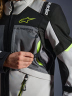 Chaqueta Alpinestars Andes V4 Drystar®, chaqueta de moto, gris oscuro, gris hielo, negro y amarillo flúor, detalle de primer plano que muestra una mano abriendo la toma de ventilación del pecho con acentos en amarillo neón y forro de malla, construcción textil duradera con la marca Alpinestars