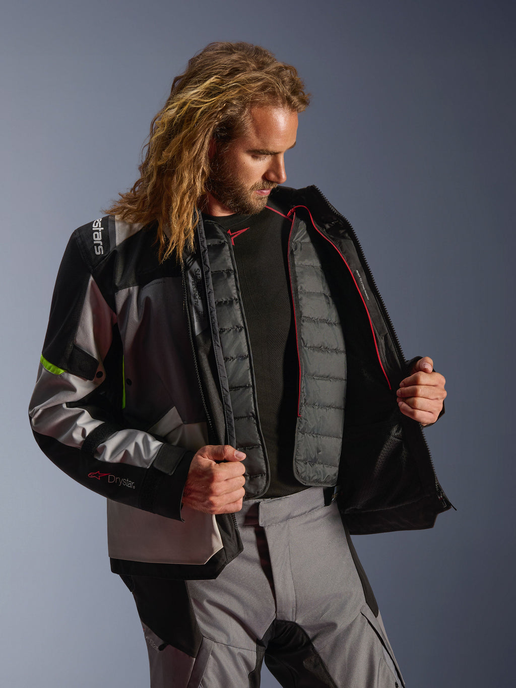 Chaqueta Alpinestars Andes V4 Drystar®, equipamiento de aventura para motociclistas en gris oscuro, gris hielo y amarillo flúor, modelo masculino mostrando el forro térmico acolchado interno, usada con pantalones de conducción a juego, diseño de alto rendimiento para todo tipo de clima