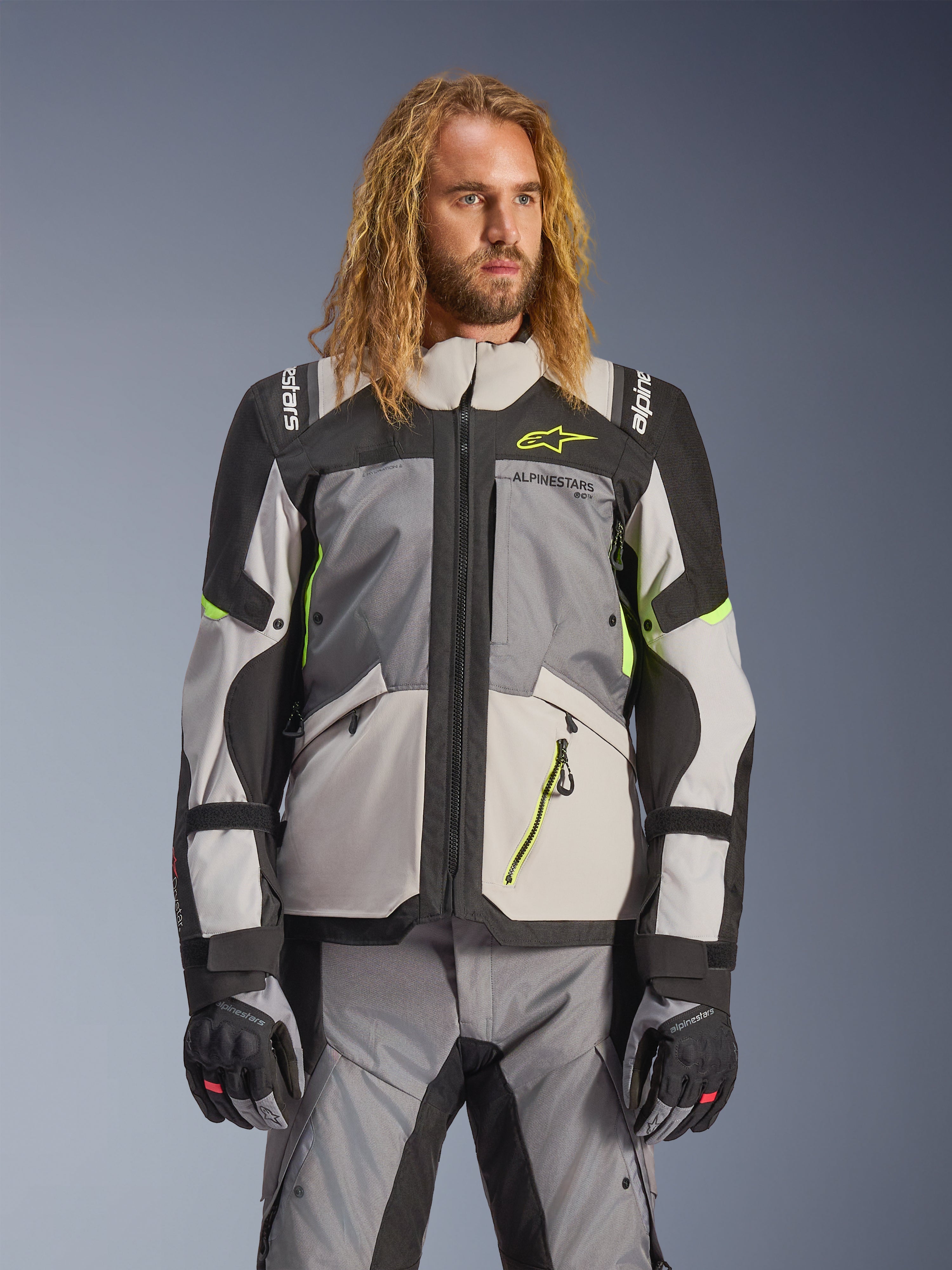 Chaqueta Alpinestars Andes V4 Drystar, chaqueta de moto para turismo de aventura, gris oscuro y gris hielo con detalles en amarillo neón, usada por un modelo con pantalones y guantes a juego, diseño textil multibolsillos para conducción en todo tipo de clima
