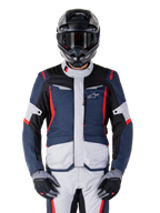 Chaqueta impermeable Alpinestars ST-1, chaqueta de turismo de aventura, azul oscuro, negro y rojo brillante, usada con casco de aventura negro y guantes grises, con paneles en gris claro y detalles rojos en hombros y cremalleras, diseño estilizado con la marca Alpinestars en el pecho, equipo impermeable duradero para motociclismo