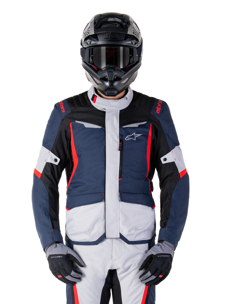 Chaqueta impermeable Alpinestars ST-1, chaqueta de turismo de aventura, azul oscuro, negro y rojo brillante, usada con casco de aventura negro y guantes grises, con paneles en gris claro y detalles rojos en hombros y cremalleras, diseño estilizado con la marca Alpinestars en el pecho, equipo impermeable duradero para motociclismo