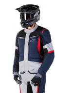 Chaqueta impermeable Alpinestars ST-1, chaqueta de moto adventure touring en azul oscuro, negro y rojo brillante, combinada con un casco negro y guantes grises, con un diseño moderno, bolsillos funcionales y el logotipo de Alpinestars.