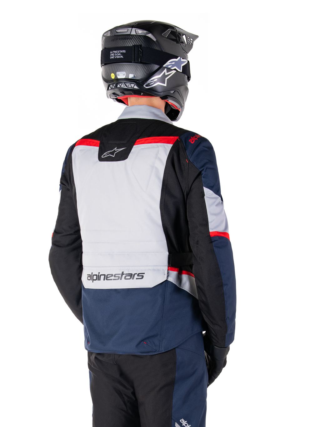 Chaqueta impermeable Alpinestars ST-1, azul oscuro, negro y rojo brillante, vista trasera de un piloto con casco de aventura negro, paneles en gris claro y marca Alpinestars, equipamiento de moto para turismo de aventura