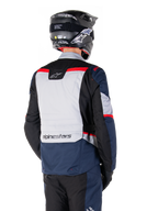 Chaqueta impermeable Alpinestars ST-1, azul oscuro, negro y rojo brillante, vista trasera de un piloto con casco de aventura negro, paneles en gris claro y marca Alpinestars, equipamiento de moto para turismo de aventura